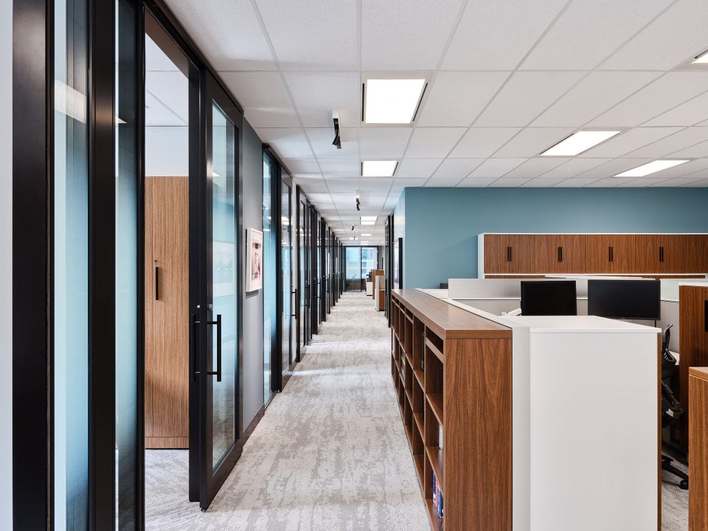 Open-plan work floor at Osler’s Ottawa office, lit by recessed ceiling troffers and adjustable track accents past glass-walled offices and a blue feature wall.