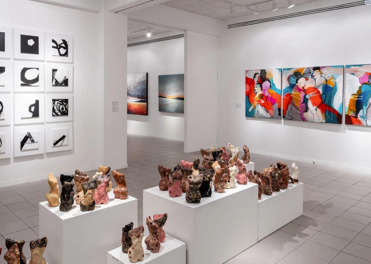 An art gallery with white plinths of small ceramic sculptures and paintings on white walls, lit by track and recessed fixtures