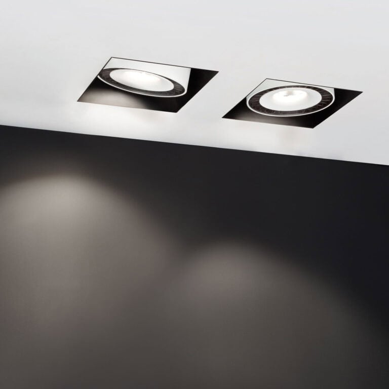 Two square trimless downlights recessed flush into a white ceiling, one angled to wash light down the dark wall below