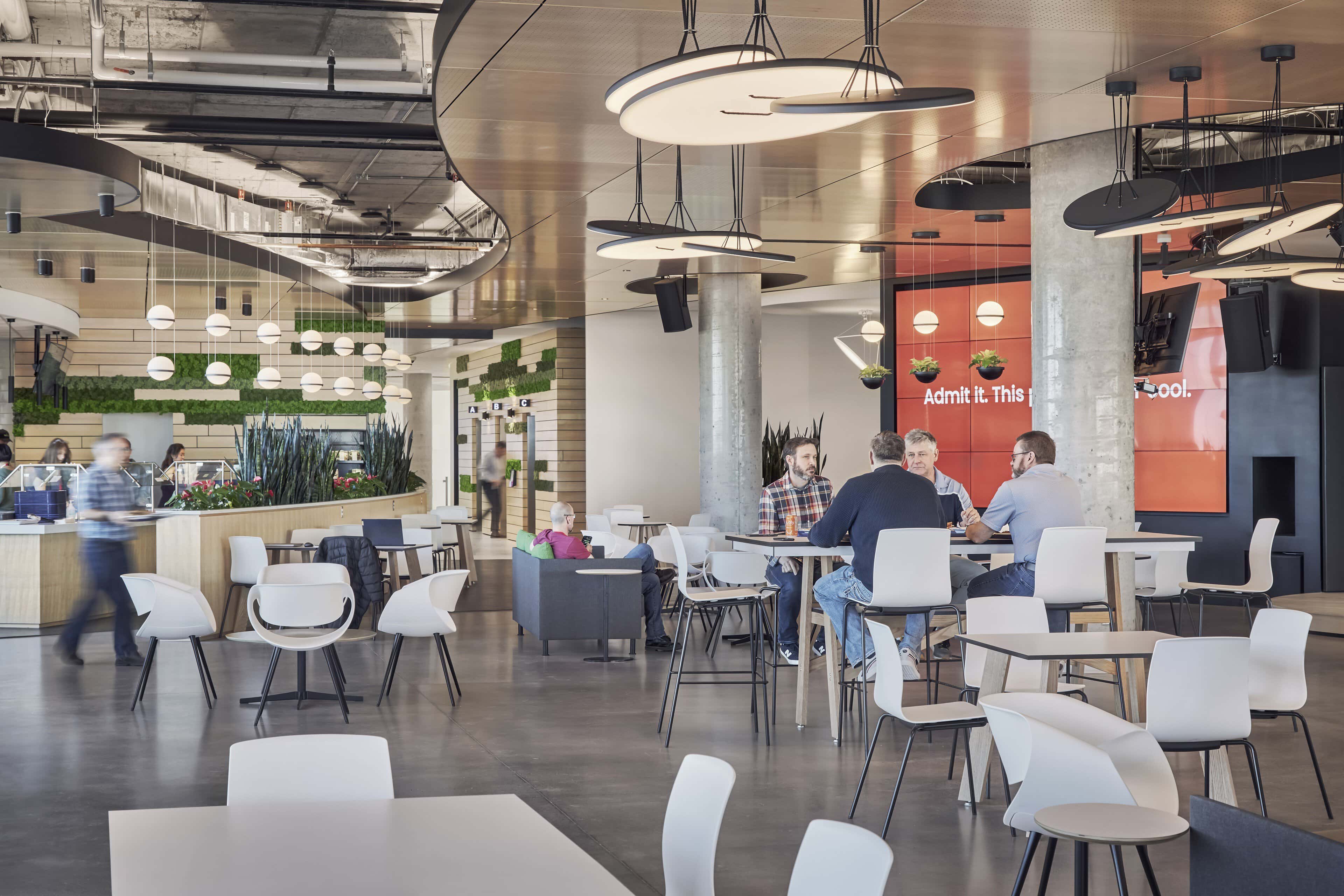 The Hive at Kinaxis headquarters in use, disc pendants and ring fixtures above the café tables with the planted green wall and servery beyond