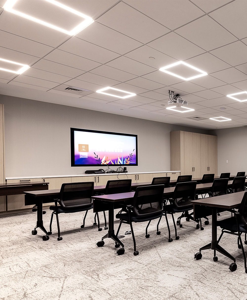 A meeting room with a suspended tile ceiling. Several Liteline EDGE panels are set into the grid, each reading as a lit rectangular outline around an unlit centre. Rows of tables on casters face a wall-mounted display.